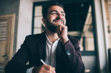 Heyecanlı, karizmatik, zarif takım elbiseli, elinde kalemle iş yerinde yüzünü çevirerek çenesine dokunan bir proje yaratan bir adamdan.