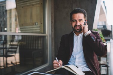 Resmi olarak İspanyol sakallı bir adam modern binanın balkonunda otururken ve akıllı telefondan konuşurken planlamacı gibi yazılar yazar ve kameraya bakar.