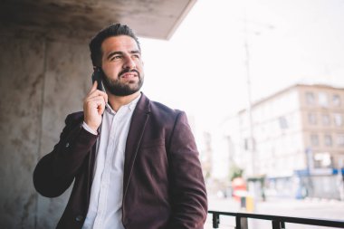 Düşünceli İspanyol takım elbiseli adam, modern şehirde çalışırken terasta akıllı telefon konuşmaları yaparken başka tarafa bakıyor ve düşünüyor.