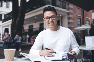 Asyalı, gözlüklü, zeki, boş not defteriyle not alan, caddedeki kafede ders çalışan ve kameraya bakan biri.