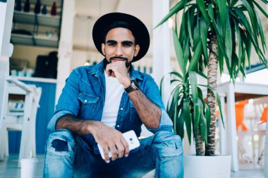 İspanyol asıllı, kendine güveni tam, modaya uygun kot giyen, cep telefonuyla takılan ve kafenin merdivenlerine yaslanmış kameraya bakan bir adamdan.