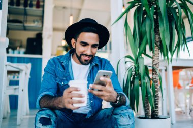 Kot pantolon ve şapka takan konsantre İspanyol erkek şehir kafesinin merdiveninde kahve içerken rahatlıyor.