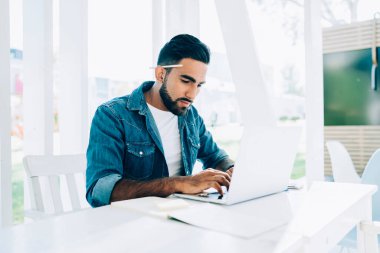 Çalışkan, düşünceli, günlük giysiler içinde, kulağının arkasında kalemle ekranda gezinen ve bilgisayarla etkileşime giren İspanyol asıllı serbest yazar açık havada beyaz terasta elinde not defteriyle otururken.