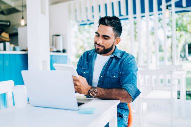 Modern açık hava kafedeki laptopta araştırma için notlar alırken şık giyinmiş, güler yüzlü etnik erkek öğrenci. 