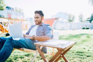 Gündelik kıyafetlerle gezen mutlu etnik adam dizüstü bilgisayarla yazı yazarak parktaki sandalyede oturarak yeni bir başlangıç yapıyor.