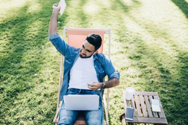 Kızgın, genç, etnik erkek, kollarını kaldırmış, sıradan bir kot gömlek ve dizüstü bilgisayarlı kot pantolon giymiş güneşli bir günde parkta, dinlenme odasında oturuyor.