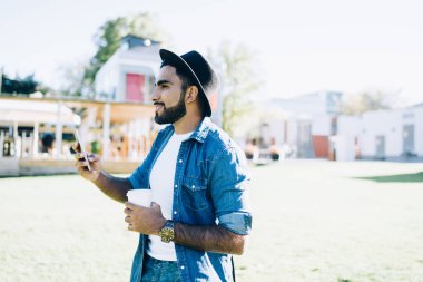 Odaklanmış gülümseyen, siyah şapkalı, beyaz gömlekli, kot elbiseli, parkta arkadaşını beklerken cep telefonuna bakan etnik bir erkek.
