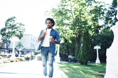 Şık şapkalı, kot pantolonlu, sakin İspanyol erkek kaldırımda gezerken cep telefonuyla güneşli parkta kahve içiyor.