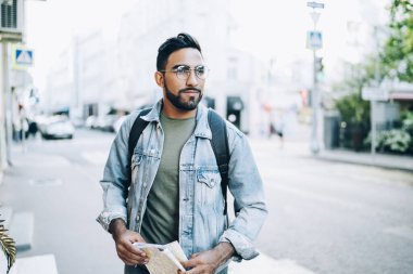 Gözlüklü, sıradan kot ceketli, sakallı, koca şehirde elinde haritayla caddede duran mutlu bir etnik erkek.