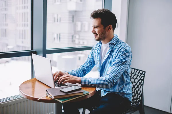 From above of confident joyful man in casual wear working on freelance project using modern laptop typing sitting at table at work space