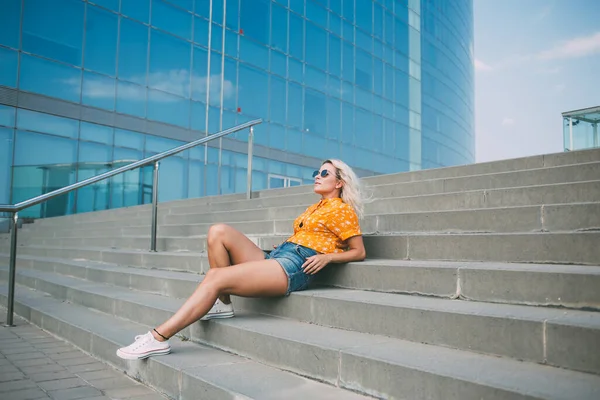 フルボディ流行の女性でカジュアル夏服上に横たわるステップと都市の通りに現代的なガラスの建物の外で休んで — ストック写真