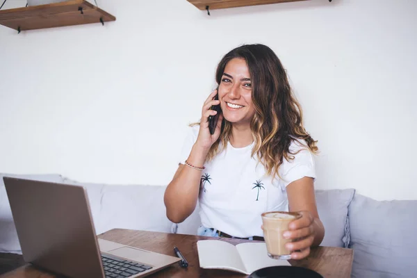 魅力的な幸せな笑顔の若い女性とコーヒーを片手にノートパソコンの前のカフェに座って電話で話す — ストック写真
