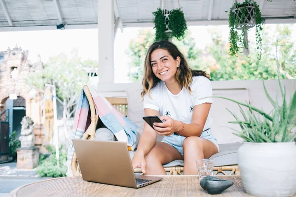 stock image Dark haired young cheerful female with toothy smile sitting on comfortable sofa near table with glass of water while typing on smartphone and using laptop