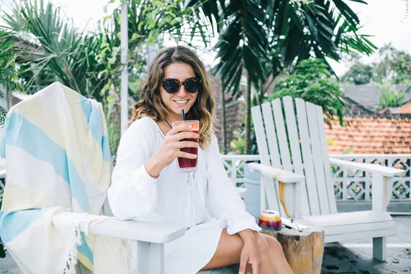 愉快地满足了这位戴着太阳镜的轻松微笑的女士 她坐在椅子上 一边享受着红色的滑滑 一边在度假咖啡馆里享用早餐 — 图库照片