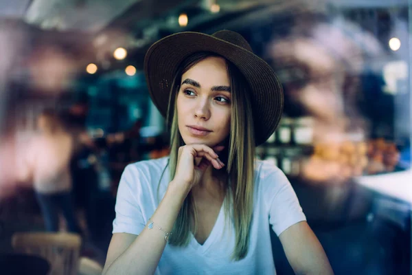 左側を見ている熱帯の帽子と白いシャツの若い女性を熟考し 背景にぼやけたカフェに座っている間に顎の近くに手を保持するガラスビューを通して — ストック写真