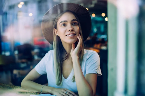 Mujer Alegre Positiva Camiseta Sombrero Sentado Cafetería Tocando Cara Mientras —  Fotos de Stock