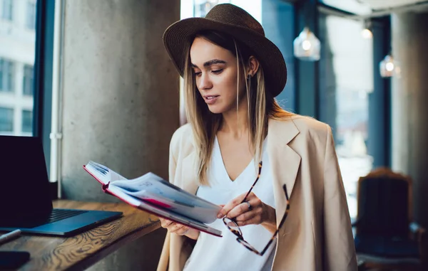 Young Woman Stylish Hat Holding Glasses Flipping Pages Notepad While — Stock Photo, Image