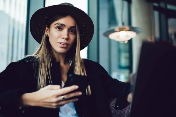 スタイリッシュな流行のスマート女性で帽子作業ノートパソコンに座ってアームチェアとサーフィン携帯電話でカメラを見て質問顔 — ストック写真