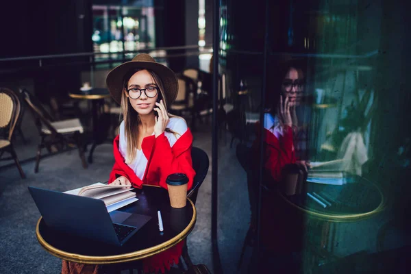 戴帽子戴眼镜 头戴五颜六色的红色毛毯的自信女人一边说着智能手机 一边坐在街边咖啡店的桌子旁 手里拿着笔记本电脑和咖啡杯 — 图库照片