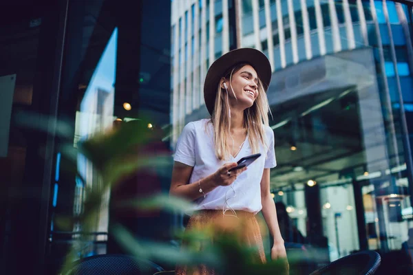 幸せな女性の帽子やイヤフォンのカジュアルな服を着て通りに立ってガラスの壁の近くに手にスマートフォンと離れて笑みを浮かべて見て — ストック写真