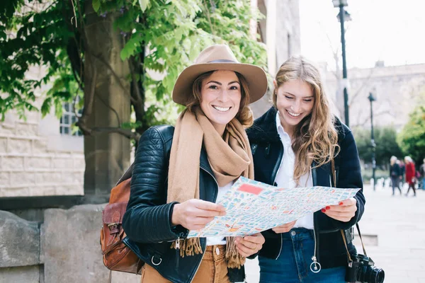 Cheerful young female tourists in stylish clothes with backpack and photo camera standing together next to old building and reading city map while walking in city