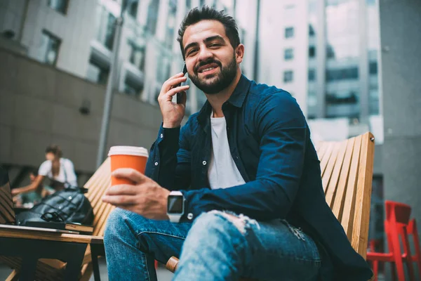 Handsome caucasian male student recreating during coffee break talking on mobile phone in roaming, portrait of young prosperous hipster guy sitting outdoors enjoying smartphone conversation