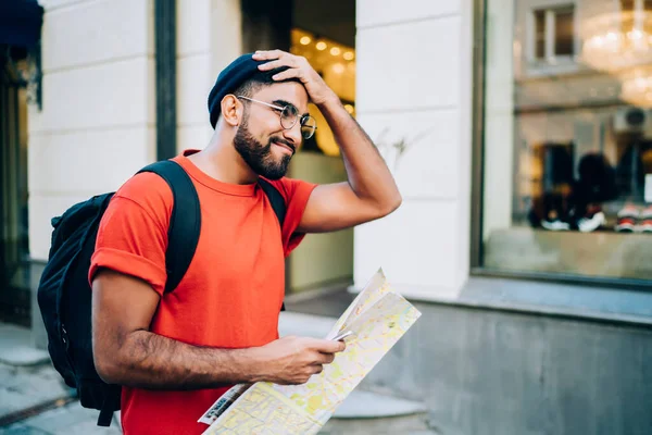 Stylish bearded ethnic male on vacation with backpack in trendy casual outfit and eyeglasses taking wrong turn while navigating with map at city street