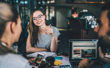Genç neşeli iş arkadaşları cep telefonlarıyla mesajlaşıp dizüstü bilgisayarlarını izliyorlar. Yaratıcı iş yerindeki renkli çıkartmalarla dolu masada oturuyorlar. 