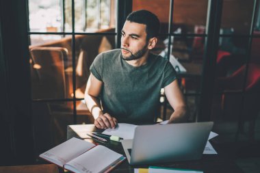 Ciddi sakallı bir adam modern dizüstü bilgisayarını karıştırıyor ve masada oturup rahat bir restoranda serbest iş yaparken gözlerini kaçırıyor.