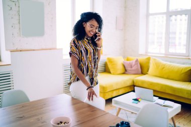 Ev kıyafetleri ve gözlükleriyle gülümseyen Afro-Amerikan genç bir kadın şık bir dairede sandalyeye yaslanırken akıllı telefondan sohbet ediyor.