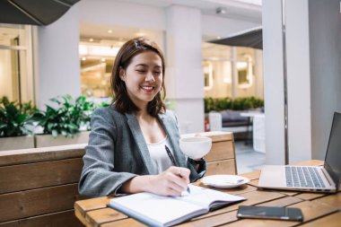 Genç gülümseyen Asyalı kadın serbest yazar not defterine not alıyor ve kafede dizüstü bilgisayarı ve akıllı telefonu ahşap masada otururken kahve içiyor. 