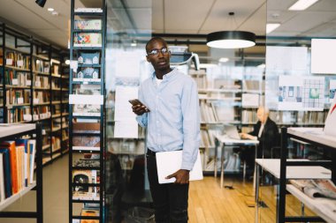 Açık tişörtlü akıllı Afro-Amerikalı adam cep telefonu ve kitaplığıyla kitapçıda beklerken bekliyor.