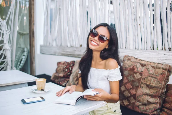 Omzunun dışında bir bluz ve güneş gözlüğü takmış, elinde kahve ve akıllı telefonla tropik bir kafede oturmuş kitap okuyan mutlu Asyalı genç bir kadın.