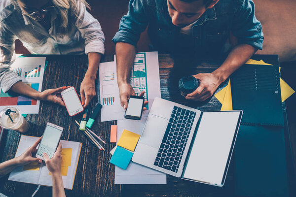 From above of crop colleagues having break during meeting and checking and sending messages on mobile phones while sitting together at table with papers and laptop with white blank screen 