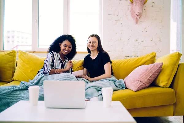 Smiling African American woman with diverse female friend sitting on yellow sofa under blue cozy plaid and watching comedy movie on laptop