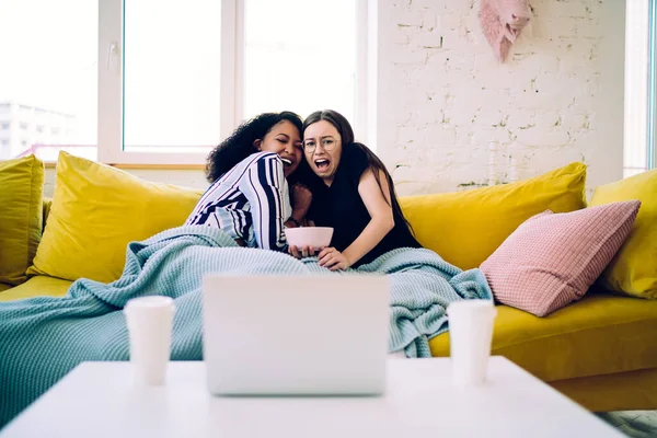 Terrified diverse female teenagers leaning against each other and screaming in fear while sitting on yellow sofa under blanket and watching scary movie on laptop