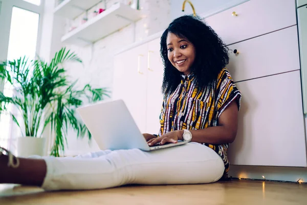 Pleasantly surprised African American female freelancer with black curly hair in colorful shirt working on portable computer while sitting on kitchen floor