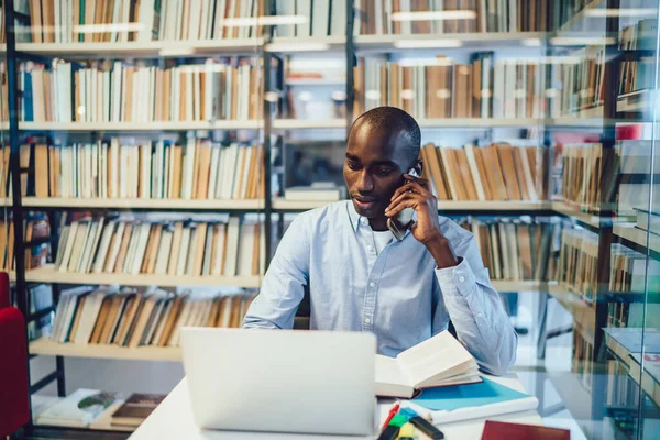 ノートパソコンを使い携帯電話で話す黒人男性に焦点を当て 大学図書館の書棚の近くで宿題をする — ストック写真