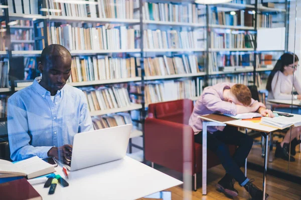 穿着正式西服 心事重重的多种族工作者坐在疲倦的同事旁边 脸朝下躺在书桌上 图书馆里放书 — 图库照片