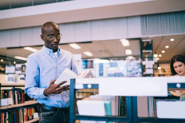 身穿衬衫和眼镜的勤勉的非洲裔美国年轻人一边翻动着几页书 一边站在现代图书馆的书架上 — 图库照片