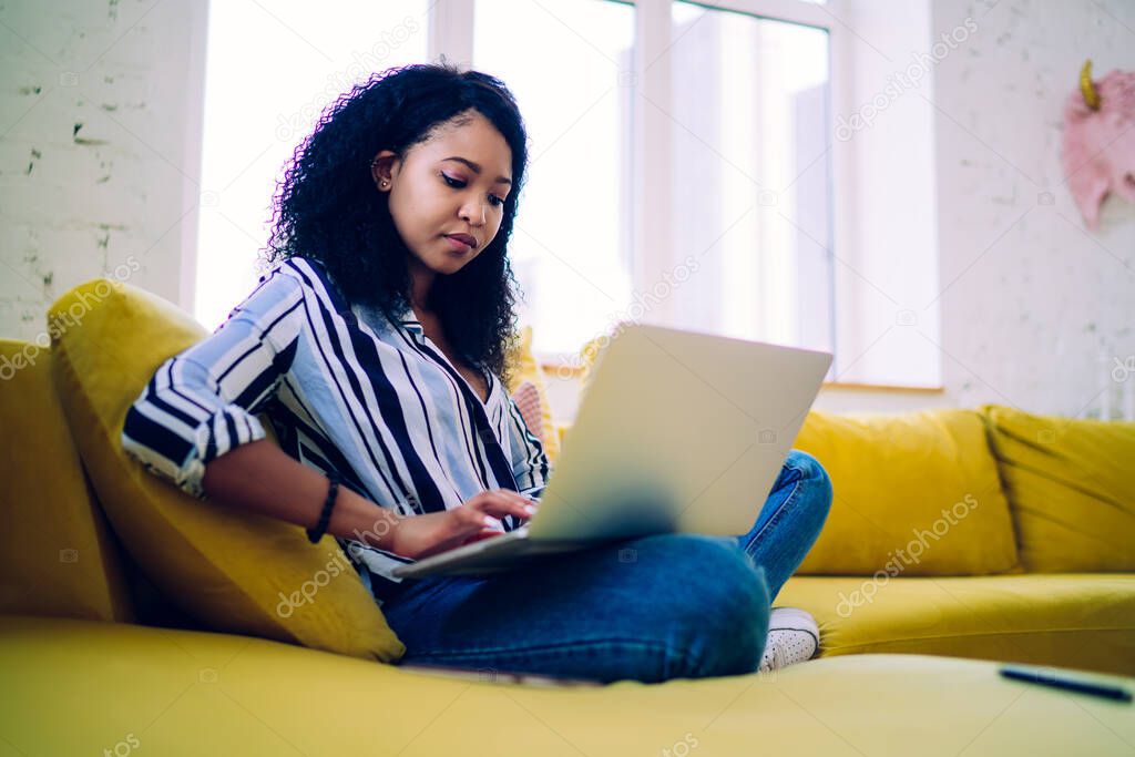 Busy and confident African American freelancer in striped shirt sitting on yellow sofa and working at home while typing message in social media