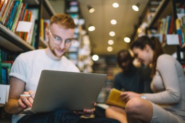 Kütüphane zemininde kitap rafları arasında milli etnik arkadaşlarla oturan genç konsantre kızıl adamın elindeki dizüstü bilgisayara odaklan.