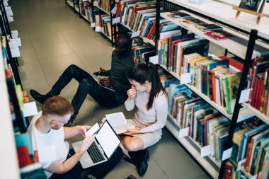 Yukarıdan bakıldığında, düşünceli, çok ırklı öğrencilerin kitaplıklar arasında birlikte projeye hazırlandıkları kitapların ve cihazların arasında oturdukları görülüyor.