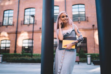 Sütunun yanında durup, şehir caddesinde hoş bir akıllı telefon konuşması yapan, gülümseyen ve yüzünü çeviren defterlerle mutlu genç bir kadın.