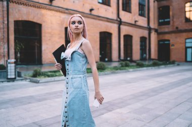 Pembe saçlı, elinde defterler ve akıllı telefonlar taşıyan ve kaldırımda yürürken yüzünü çeviren modaya uygun genç bir bayan.