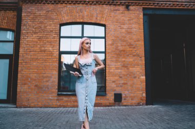 Elinde not defterleri olan modaya uyan genç bir kadın şehir caddesinde tuğla binanın önünde dururken akıllı telefonu cebine koyuyor.