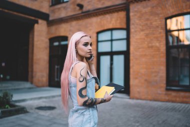 Kolu dövmeli, uzun pembe saçlı, kot elbiseli, cep telefonuyla konuşan, elinde sarı kitapla şık bir kadın. 