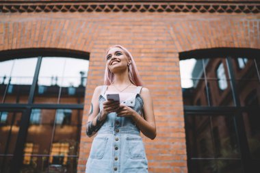 Dövmeli, uzun pembe saçlı, kot elbiseli, caddede cep telefonunda sörf yapan modern, neşeli bir bayan.