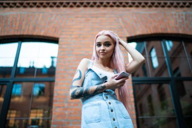 Düşük açılı, güler yüzlü, yılan dövmeli, uzun pembe saçlı, kot elbiseli, sokakta cep telefonuyla mesajlaşan bir kadın.