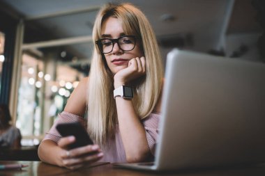 Akıllı telefondan mesaj okuyan genç ve düşünceli bir kız öğrencinin laptopuyla masada oturması ve kafede sınava hazırlanması.
